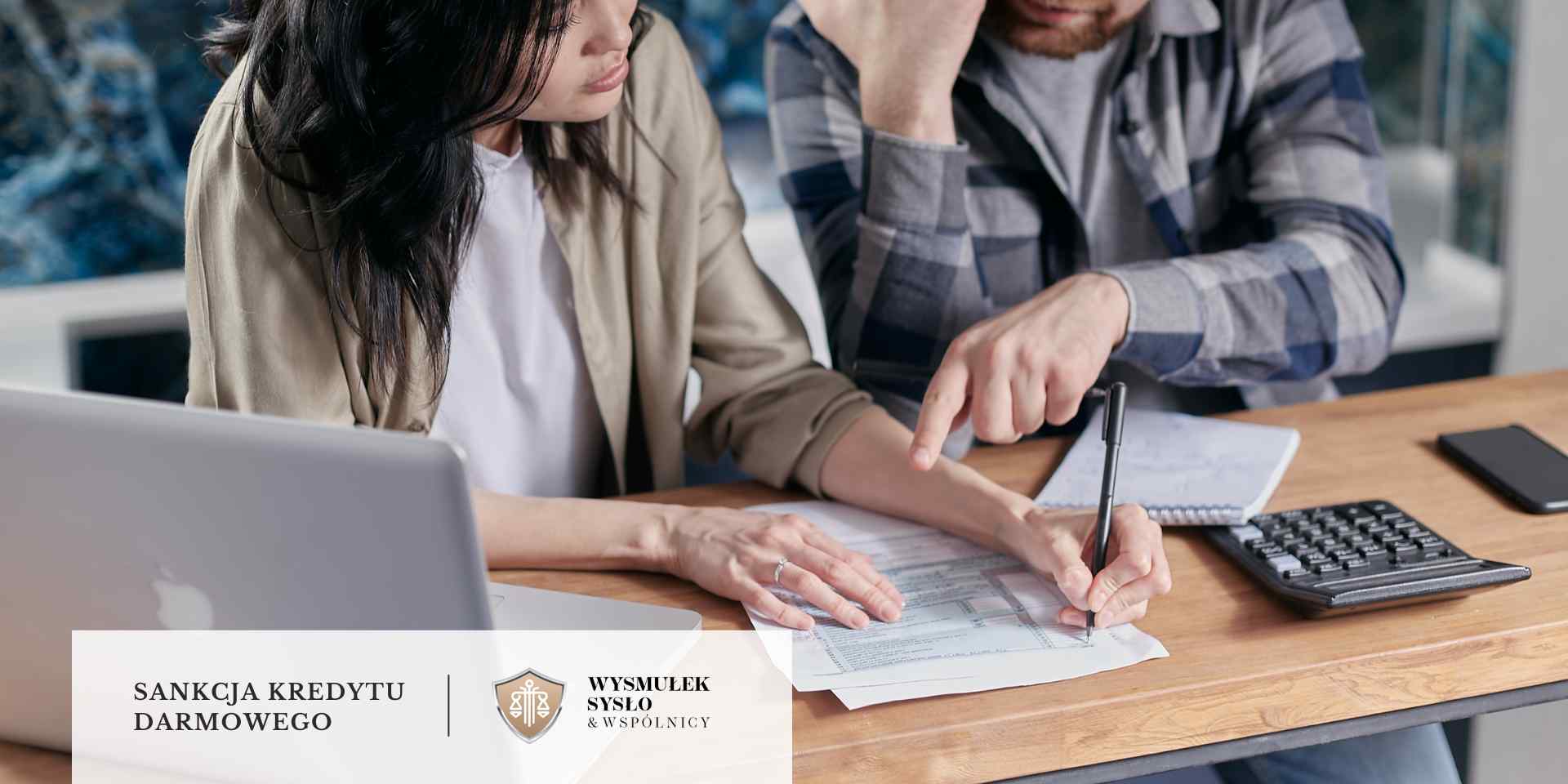 
Na drewnianym stole para przegląda dokumenty i podpisuje je, w tle widoczny jest laptop i kalkulator