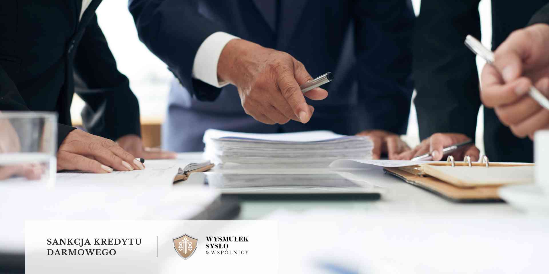 Grupa prawników w ciemnych garniturach pochylona nad stosem dokumentów na białym stole.