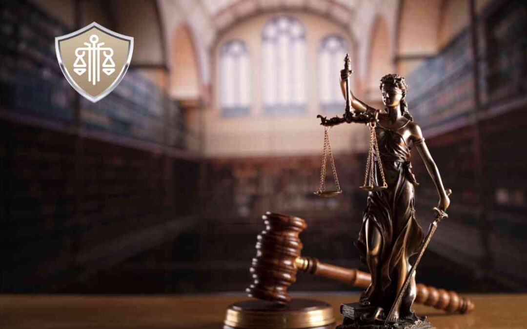 Statue of Lady Justice holding scales and a sword in a courtroom setting, with a gavel on the desk and a blurred courthouse backdrop.